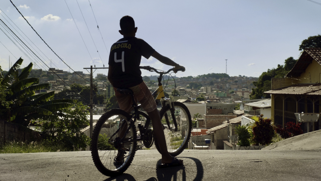 Fotografia de uma cena do filme Marte um, de Gabriel Martins. MG, 2022, FICÇÃO, 115 min. De costas, jovem negro em uma bicicleta no alto de uma ladeira. Ao longe, um morro. À esquerda, plantas e bananeiras e, à direita, diversas casas. Ele usa camisa esportiva preta com o número 4 estampado nas costas, bermuda rosa e chinelo preto. Fios elétricos cortam o céu azul.