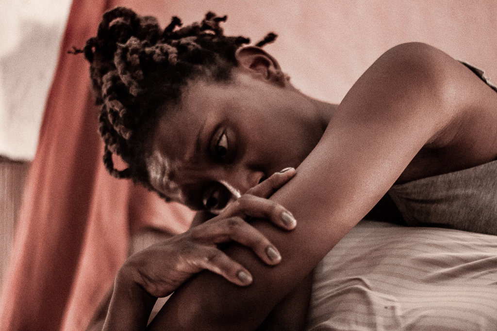 Fotografia de uma cena do filme. Uma pessoa negra deitada, com a cabeça voltada para a esquerda, tem os cabelos pretos com tranças avermelhadas presas num coque. Tem a testa franzida, as sobrancelhas finas, arqueadas, olha desolada para baixo. A mão direita está sobre o braço esquerdo estendido para baixo. De camiseta regata cinza mescla, está deitada sobre algo coberto por um lençol bege de listras rosadas. Atrás, desfocado um tecido parte rosa, parte vermelha e outra branca.