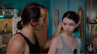 Fotografia de uma cena do filme A filha do palhaço, de Pedro Diógenes. CE, 2022, FICÇÃO, 104 min. Em uma cozinha, em pé, pai e filha lado a lado. À esquerda, Renato, homem branco de rabo de cavalo e camiseta regata preta, toca o cabelo de Joana, uma adolescente branca de rabo de cavalo e camiseta regata cinza. Ele esboça um leve sorriso, enquanto ela tem uma expressão entristecida.