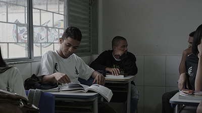 Fotografia de uma cena do filme Aulas que matei, de Amanda Devulsky e Pedro B. Garcia. DF, 2018, FICÇÃO, 23 min. Em uma sala de aula, estudantes do Ensino Médio sentados em duas fileiras. Um dos garotos, sentado numa carteira ao lado de uma janela de vidro, folheia um livro. Atrás dele, na última carteira da sala, um garoto conversa com outro estudante da fileira da direita.