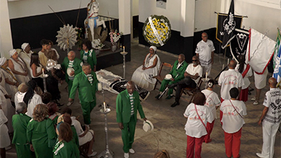 Fotografia de uma cena do filme Belos Carnavais, de Thiago B. Mendonça. SP, 2020, FICÇÃO, 16 min. Um velório. Em volta do caixão com o corpo de um homem, integrantes de uma escola de samba. À esquerda, em pé, mulheres com roupas do candomblé e pessoas de uniforme verde e branco. À direita, em pé, mulheres de camiseta branca e calça vermelha. Sentadas perto do caixão, três pessoas. Atrás delas, estandartes pretos com o nome Vai-Vai em letras brancas e uma coroa de flores. Sobre um pequeno palco ao fundo do salão, uma imagem, do tamanho de um adulto, de São Jorge montado em seu cavalo branco.