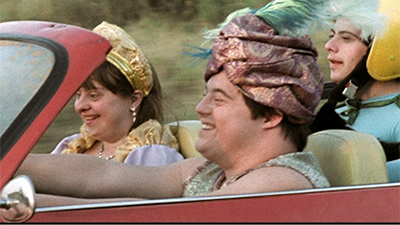 Fotografia de uma cena do filme Colegas, de Marcelo Galvão. SP, 2012, FICÇÃO, 100 min. Três pessoas brancas em um carro conversível vermelho, direcionado para a esquerda. No volante, o homem usa um turbante roxo com uma pena verde. No rosto arredondado, ele tem os olhos puxados, quase fechados e um sorriso largo. Usa uma camiseta regata, verde e rosa e está com os braços estendidos, no volante. Ao seu lado, a mulher de cabelos lisos, castanhos claros até o ombros e franja, usa uma faixa dourada na cabeça. Com os miúdos olhos voltados para baixo, ela sorri levemente. Usa brinco e colar com pérolas e tem as mangas bufantes do vestido, douradas. No banco de trás, o outro homem está com um capacete amarelo, tem os pequenos olhos puxados voltados para frente e sorri discreto, usa uma blusa de lycra verde claro, com uma fita verde escuro na gola. No fundo distorcido, a vegetação sugere que o carro está em movimento.