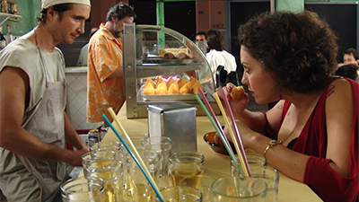 Fotografia de uma cena do filme Estômago, de Marcos Jorge. PR, 2007, FICÇÃO, 112 min. Sobre o balcão de uma lanchonete, copos de vidro com canudos, um porta-guardanapo de alumínio e uma estufa repleta de coxinhas. Do lado de dentro, um homem branco com chapéu de cozinheiro, camiseta regata e avental brancos. Do lado de fora, uma mulher branca, de cabelos castanhos e ondulados até o pescoço, usa uma blusa vermelha decotada. Eles se olham fixamente enquanto ela cheira um salgado já mordido.