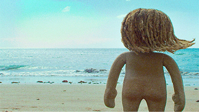 Fotografia de uma cena do filme Guaxuma, de Nara Normande. PE, 2018, ANIMAÇÃO/FICÇÃO, 15 min. Dia de céu azul. Em uma praia, um boneco de areia, de costas, mirando o mar. Ele tem cabelos dreadlocks que esvoaçam para a direita.