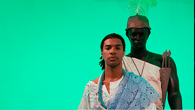 Fotografia de uma cena do filme Joãosinho da Goméa, O Rei do Candomblé, de Janaína Oliveira e Rodrigo Dutra. RJ, 2019, DOCUMENTÁRIO, 15 min. Contra fundo verde, de frente e sério, Joãosinho da Goméa nos encara. Ele é um homem negro de pele não retinta. O cabelo cacheado está preso em um rabo de cavalo que vai até o pescoço. Usa uma bata branca adornada com linhas azuis. Sobre o ombro esquerdo, uma manta azul de Richelieu. No pescoço, um colar branco e outro azul. Atrás de Joãosinho, um manequim negro de pele retinta representa Oxóssi. O orixá, mais alto que o Rei do Candomblé, também nos encara. Ele usa um capacete metálico com um ramo verde e longo ao centro e uma túnica branca amarrada sobre o ombro direito. Sobre o ombro esquerdo, Oxóssi carrega três flechas dentro de uma bolsa cônica de metal.