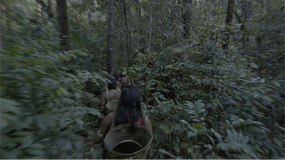 Fotografia de uma cena do filme Mãri Hi – A Árvore do Sonho, de Morzaniel Iramari. RR, 2023, FICÇÃO, 18 min. Na cena desfocada, uma fila de indígenas de costas, carrega cestos nas costas, adentram a floresta densa e verde. A indígena do fim da fila tem longos cabelos lisos e pretos.