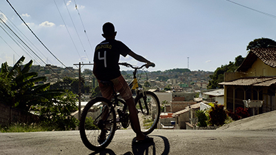 Fotografia de uma cena do filme Marte um, de Gabriel Martins. MG, 2022, FICÇÃO, 115 min. De costas, jovem negro em uma bicicleta no alto de uma ladeira. Ao longe, um morro. À esquerda, plantas e bananeiras e, à direita, diversas casas. Ele usa camisa esportiva preta com o número 4 estampado nas costas, bermuda rosa e chinelo preto. Fios elétricos cortam o céu azul.