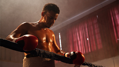Fotografia de uma cena do filme Mirador, de Bruno Costa. PR, 2021, FICÇÃO, 95 min. Em uma sala com janelas cobertas por cortinas vermelhas, Maycon, um homem negro não retinto de cabelos cacheados curtos com o corpo suado, luvas de boxe vermelhas e calção branco. Sério, de olhos fechados e cabeça baixa, se apoia na corda preta de um ringue.