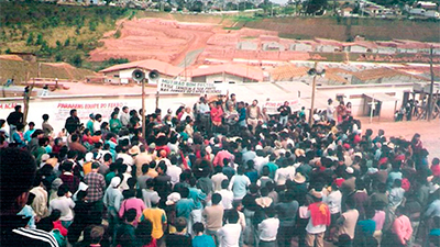 Fotografia de uma cena do filme Mutirão, de Lincoln Péricles. SP, 2022, DOCUMENTÁRIO, 10 min. Em uma rua de terra batida de uma comunidade, centenas de pessoas em pé aglomeradas em frente a um palanque ocupado por várias outras pessoas. Atrás, três faixas com texto desfocado estendidas ao longo de um muro branco que separa um terreno com vegetação à esquerda, uma área de barro e, à direita, casas de arquitetura semelhante, alinhadas, com telhas vermelhas e paredes brancas.