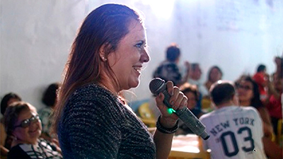 Fotografia de uma cena do filme Noite de seresta, de Muniz Filho e Sávio Fernandes. CE, 2020, DOCUMENTÁRIO, 19 min. Em um salão, de perfil, uma mulher branca com cabelos castanhos lisos longos e suados usa camisa preta com finas listras prateadas. Com um sorriso largo, segura um microfone brilhante prateado. Ao fundo, pessoas desfocadas sentadas ao redor de mesas plásticas, amarelas. À esquerda, uma senhora de óculos de grau, olha sorridente para a mulher.