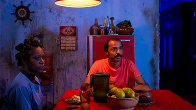 Fotografia de uma cena do filme O ovo, de Rayane Teles. BA, 2022, FICÇÃO, 23 min. Em uma cozinha iluminada por luz lilás, uma mulher e um homem negros sentados em volta de uma mesa vermelha quadrada com prato, copo, jarra de suco, vaso de barro com laranjas e panela de barro tampada. À esquerda, a mulher usa penteado com diversos coques e camisa roxa. De frente para nós, o homem tem cabelos curtos e bigode pretos e usa camisa vermelha. Ao fundo, na parede com manchas pretas de mofo, um leme de barco e calendário pendurados. Atrás do homem, uma geladeira vermelha com moringa, garrafas e cesta de fruta. Eles olham sérios para a diagonal direita.