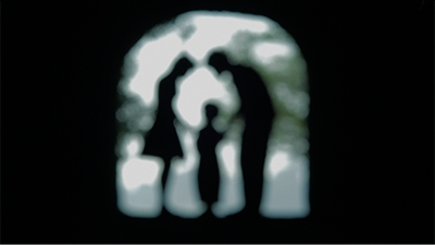 Fotografia de uma cena do filme Outubro acabou, Karen Akerman e Miguel Seabra Lopes. BR/PT, 2015, DOC/DRAMA, 24 min. Silhuetas desfocadas de três pessoas em pé sob um arco. A pessoa à esquerda usa saia e as outras, camisa e calça. A do centro é bem mais baixa e as outras duas se inclinam quase encostando as cabeças e as mãos na do meio.