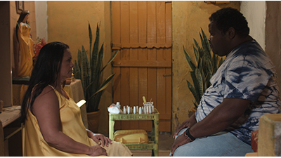 Fotografia de uma cena do filme Quebra panela, de Rafael Anaroli. PE, 2022, FICÇÃO, 17 min. Em uma sala de paredes amarelas, uma mulher e um homem estão sentados frente a frente. Sérios, se encaram. São negros e gordos. Ela tem cabelos pretos longos lisos e usa vestido amarelo. Ele tem cabelos curtos cacheados pretos. Está com uma camisa de tie-dye azul e branca e calça jeans. Entre eles, uma pequena estante de plástico verde, com esmaltes, algodão e lixas. Mais atrás, à esquerda, sobre uma mesa, está uma escultura de uma mulher branca, com cabelos pretos e vestido longo amarelo. Ao fundo, uma porta de madeira amarelada e, de cada lado, um vaso com palmas da planta espada de São Jorge.