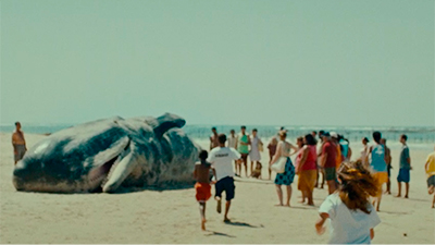 Fotografia de uma cena do filme Sem coração, de Nara Normande e Tião. PE, 2023, FICÇÃO, 91 min. Na praia, pessoas olham uma baleia encalhada na areia. Algumas pessoas correm em direção ao grupo. Estão com blusas e bermudas. O céu está azul e o mar longe. A faixa de areia se mostra extensa.