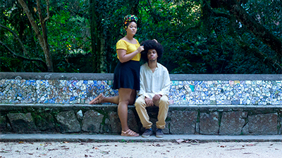 Fotografia de uma cena do filme Travessia, de Safira Moreira. BA, 2018, DOCUMENTÁRIO, 5 min. Uma mulher e um homem negros em ambiente com árvores e vegetação densa ao fundo e um grande banco revestido por mosaicos de azulejos azuis e brancos. Ela usa turbante preto com estampa colorida, camisa amarela, saia preta e sandália bege. Ele tem cabelos black power preto e veste bata branca de mangas longas, calça bege e tênis pretos. A mulher, em pé e com a perna esquerda apoiada no banco, ajeita os cabelos do homem sentado com as mãos entre as pernas. Olham sérios para a frente.