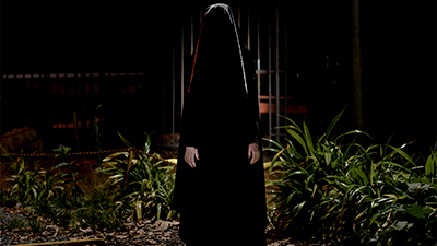 Fotografia de uma cena do filme A triste figura, de Calebe Lopes. BA, 2018, FICÇÃO, 18 min. Na noite escura, uma pessoa com véu preto sobre a cabeça e uma túnica larga embaixo, também preta, de manga comprida, tem as mãos brancas, ao lado das coxas. Uma luz fria, por trás contorna a cabeça e o véu até abaixo dos ombros e as plantas, nas laterais da estranha encapuzada. Ao fundo, um portão e grades de ferro estão na penumbra.