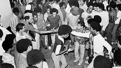 Fotografia de uma cena do filme Black Rio! Black Power!, de Emílio Domingos, RJ, 2023, Documentário, 75 min. Foto em preto e branco entre as décadas de 70 e 80. Pessoas negras com cabelos crespos em diversos tamanhos reunidas em um baile. Ao centro um jovem dança, usa camisa branca de manga 3/4, colete preto e calça branca. Próximo a ele dois homens se cumprimentam, um deles, usa camisa branca de mangas longas e cachecol quadriculado. Ao redor deles pessoas conversam e sorriem.