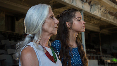 Fotografia de uma cena do filme Guerra de algodão, de Marília Hughes e Cláudio Marques - BA, 2019, FICÇÃO, 82 min. Duas mulheres brancas, de perfil direito, do busto para cima. A primeira, à esquerda, Maria, olha fixo à frente, tem cabelos lisos grisalhos ao longo das costas, presos para trás, nariz afilado e pele enrugada. Usa roupa clara, colar vermelho e verde. Ao lado, Dora, olhar decidido, nariz e queixo empinado, cabelos lisos longos, castanhos claro, com trança presa para trás e o restante solto, e pele lisa cintilante. Usa roupa de mangas, azul escura com detalhes triangular branco. O fundo desfocado, é um ambiente pouco iluminado com arquitetura antiga descascada, em tons pasteis, com poltronas enfileiradas, janela escura e teto rebaixado.