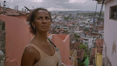 Fotografia de uma cena do filme Inabitável, de Matheus Farias e Enock Carvalho. PE, 2020, FICÇÃO, 19 min., Cor, Digital DCP. A atriz Luciana Souza, mulher negra, cabelos crespos cacheados, castanhos claros, presos para trás, de perfil direito, do busto para cima. Encontra-se em estreita ladeira, composta de várias casas coloridas e poucas fiações elétricas em declive, onde do alto temos em horizonte, em plano desfocado, a paisagem urbana das edificações mistas que se fundem com o panorama acinzentado da serra e do céu. A mulher tem a cabeça levemente erguida, olhar distante e para o alto, sobrancelhas afinadas, cenho levemente franzido, boca suavemente curvada para baixo, usa pequenos brincos claros, camisa regata bege, bolsa branca de curtas alças escuras sobre o ombro esquerdo.