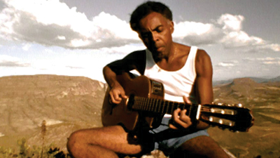 Fotografia de uma cena do filme Tempo rei, de Lula Buarque de Holanda, Andrucha Waddington e Breno Silveira. RJ, 1996, DOCUMENTÁRIO, 107 min., Cor, H264. O jovem cantor Gilberto Gil, homem negro de pele escura, cabelos pretos crespos, em curto black, mais raspados nas laterais, sobrancelhas volumosas pretas, cenho franzido, nariz arredondado de ponta mais fina, orelhas pequenas, lábios grossos em meio bico. Usa camisa regata branca, short azul curto, sentado no topo de um morro, tem a cabeça levemente inclinada para sua direita, olhar fixo para a própria mão esquerda que dedilha no braço do violão. Ao fundo, do cenário, vários morros esverdeados que contrastam com o céu azul claro com densas nuvens brancas.