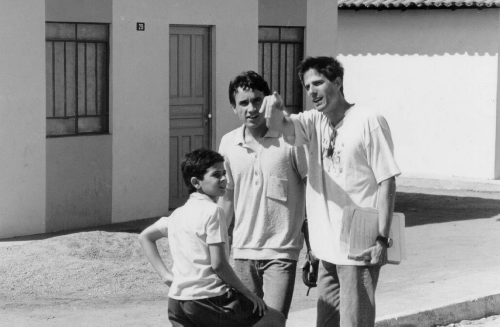 Fotografia em preto e branco de uma cena do filme Central Conquista Brasil - Os 7 Dias da Criação, de Esmon Primo. Numa manhã de sol, dois jovens e um menino, em frente a uma casa. Os três são brancos e tem cabelos curtos escuros. O mais alto, à direita, tem cabelos lisos, usa uma bata clara, um óculos escuros pendurados na bata, está de calça, segura papéis ao lado corpo e aponta para frente. À esquerda deste, o outro é um pouco menor, tem os cabelos ondulados, olha para nós, está com uma camisa pólo clara, calça e segura um cigarro na mão esquerda. Logo ao lado dele, o menino, de costas para nós, olha para a direita, com as sobrancelhas contraídas. Está com camisa de botão, de manga curta, branca e um shorts preto. Está com a mão esquerda na cintura e a outra apoiada na perna.