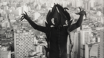 Fotografia em preto e branco de uma cena do filme. Do alto, diante dos edifícios da cidade de São Paulo ocupando todos os espaços, um demônio, de costas para nós. Na contraluz, sombreado, ele tem na cabeça adereço com pontas emendadas, que desce até abaixo da cintura, uma de cada lado. Usa uma roupa justa com uma trança longa na linha da coluna e as mãos abertas além da cabeça, tem os dedos alongados com pontas finas voltados para a cidade.