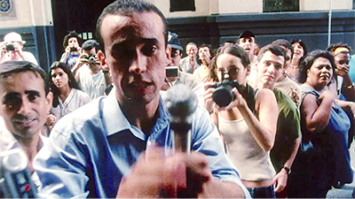 Fotografia de uma cena do filme. Em frente a um prédio antigo, um grupo grande de pessoas, com câmeras e celulares, estão direcionadas e olham um entrevistador aqui na frente. A maioria das pessoas são brancas e duas mulheres à esquerda são negras, uma delas tapa a boca com a mão. Ele é branco, magro, tem os cabelos lisos, pretos, com entradas na frente, usa uma camisa social azul clara e segura um microfone direcionado a alguém. Ele tem a testa contraída e o olho à esquerda sombreado.