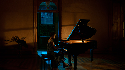 Fotografia de uma cena do filme. Na penumbra de uma grande sala, uma mulher negra, de cabelos crespos curtos, está sentada, centrada, de perfil direito, com as mãos nas teclas, diante de um piano de tampo aberto. Ela esta de vestido longo e salto. Do piano uma luz amarelada desenha sombras na parede atrás dela, que da porta no meio, verde escura, mostra pela alta janela, o escuro da noite e uma fraca luz que marca de azul o chão próximo a ela.