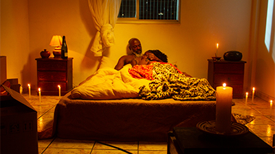 Fotografia de uma cena do filme. Num quarto, deitados numa cama de casal, Zé e uma mulher. Ele é um homem negro, de meia-idade, calvo, de barba grisalha. Está recostado na parede. Em sei peito, uma mulher de cabelos encaracolados, está deitada abraçada a ele. Estão sob cobertores e lençóis branco, vermelho e de oncinha. Acima da cabeceira, uma janela com as cortinas brancas, amarradas. De cada lado da cama, uma mesinha, à esquerda, um pote, um abajur e uma garrafa de vinho, na outra uma vela e um aparelho de som. No chão várias velas acesas e uma de sete dias, sobre um móvel, aqui na frente à direita.